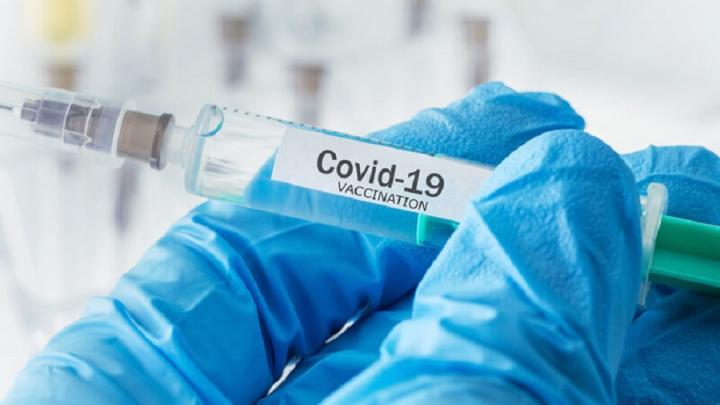 Photograph of a hand wearing a blue glove, holding a syringe labelled COVID-19 vaccination