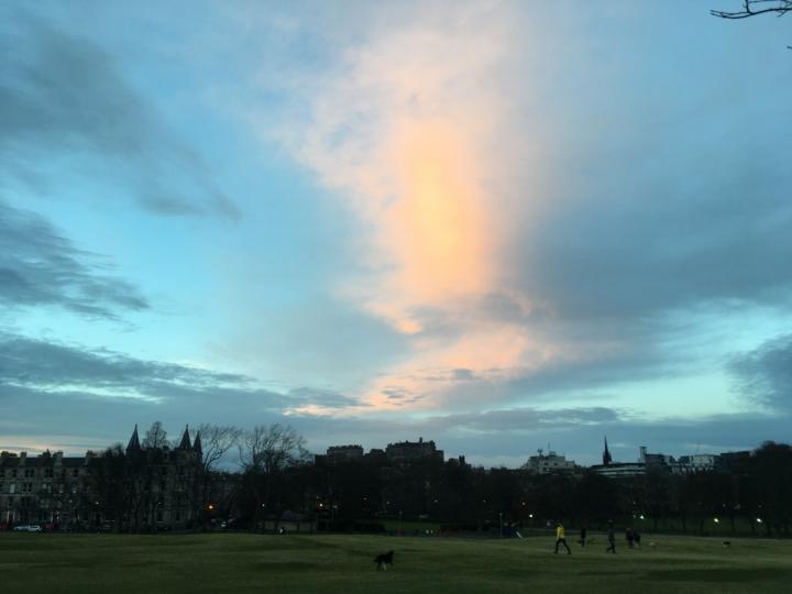 Photograph of the Meadows with Edinburgh Castle in the distance, overhead there are clouds with white, grey and pink in them.