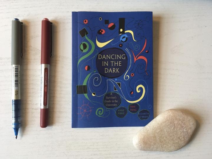 A photograph of 'Dancing in the Dark' book cover. The book is lying on a desk. The book is dark blue with green, yellow and red doodles on the cover. Beside the book is a blue and red pen. A cream stone is lying on the bottom right hand corner of the book. 