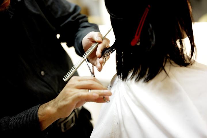 Photograph of a woman sitting in a chair getting her haircut. Behind her stands the hairdresser holding a comb and a pair of scissors
