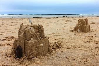 Photograph of two sandcastles on a beach, in the distance the sea meets the sand and overhead there is blue sky. 