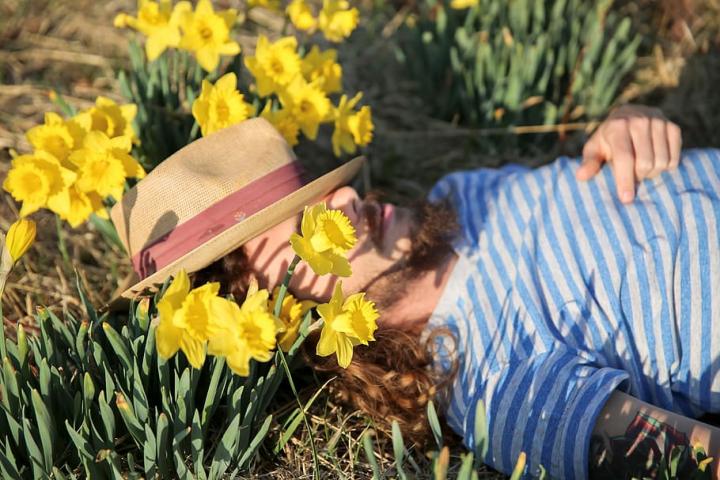 Photograph of a person lying in the grass, amongst daffodils with a summer hat covering their eyes. 