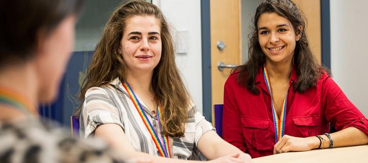 Two young women of different ethnic backgrounds sitting at a table, listening to a third woman and wearing LGBT+ lanyards