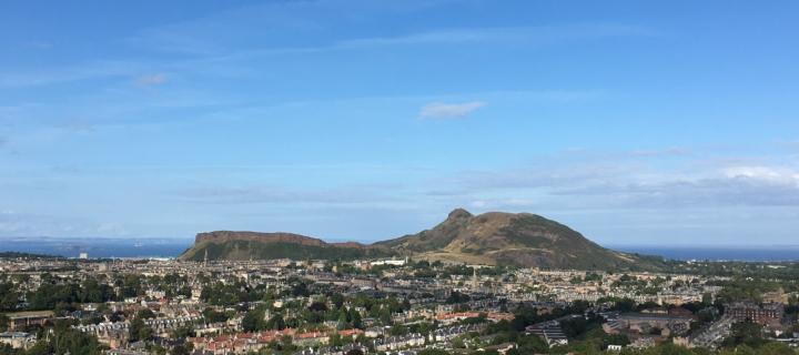 A photograph of Edinburgh with Arthur's Seat in the background. There are blue skies overhead and in the distance is the Firth o