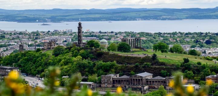 A photograph of Calton Hill in Edinburgh. In the distance you go see buildings and houses and beyond the Firth of Forth. 