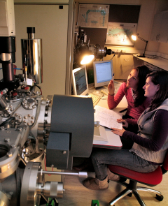 Two female scientists seated looking at computer monitors and surrounded by scientific equipment