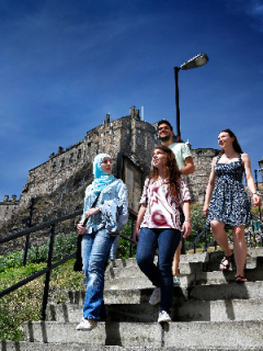 One male and three female students outdoors walking down steps with Edinburgh castle in the background