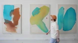 A person walks in front of three paintings at the ECA Graduate Show exhibition