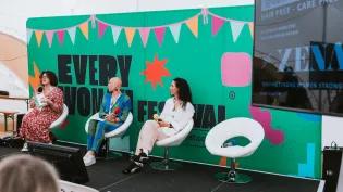 Three people sit on chairs on a stage in front of a large green banner, displaying 'Everywoman' as part of a panel discussion for Everywoman Festival