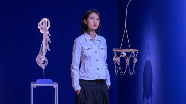 An artist stands in front of their exhibition and holds a piece featuring a woven basket with clay faces.