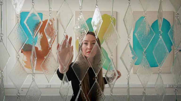 An ECA graduate approaches a sculpture that they designed made of glass and steel.