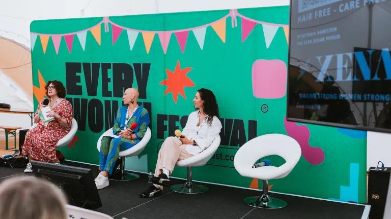Three people sit on chairs on a stage in front of a large green banner, displaying 'Everywoman' as part of a panel discussion for Everywoman Festival