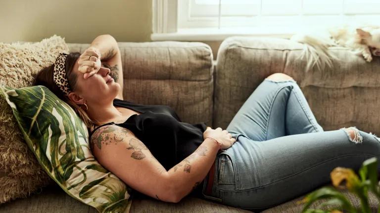 Young woman suffering from stomach cramps on the sofa at home, with hand over her face in pain