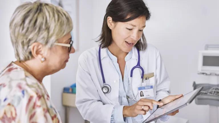 Female doctor explaining electronic test results to female patient at clinic. They are sitting in examination room and the doctor has an ipad. The doctor is wearing a white coat and has a stethoscope around her neck. The patient is wearing a floral shirt and glasses.
