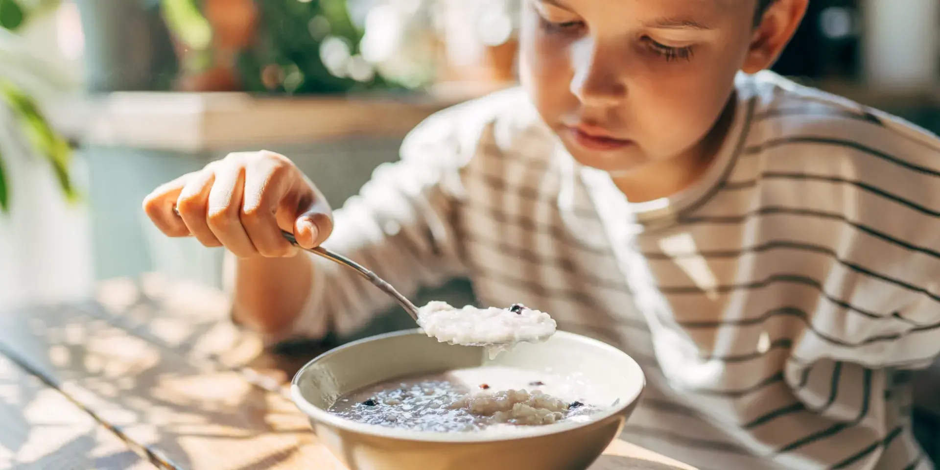 Young boy eating porridge at table near a window 