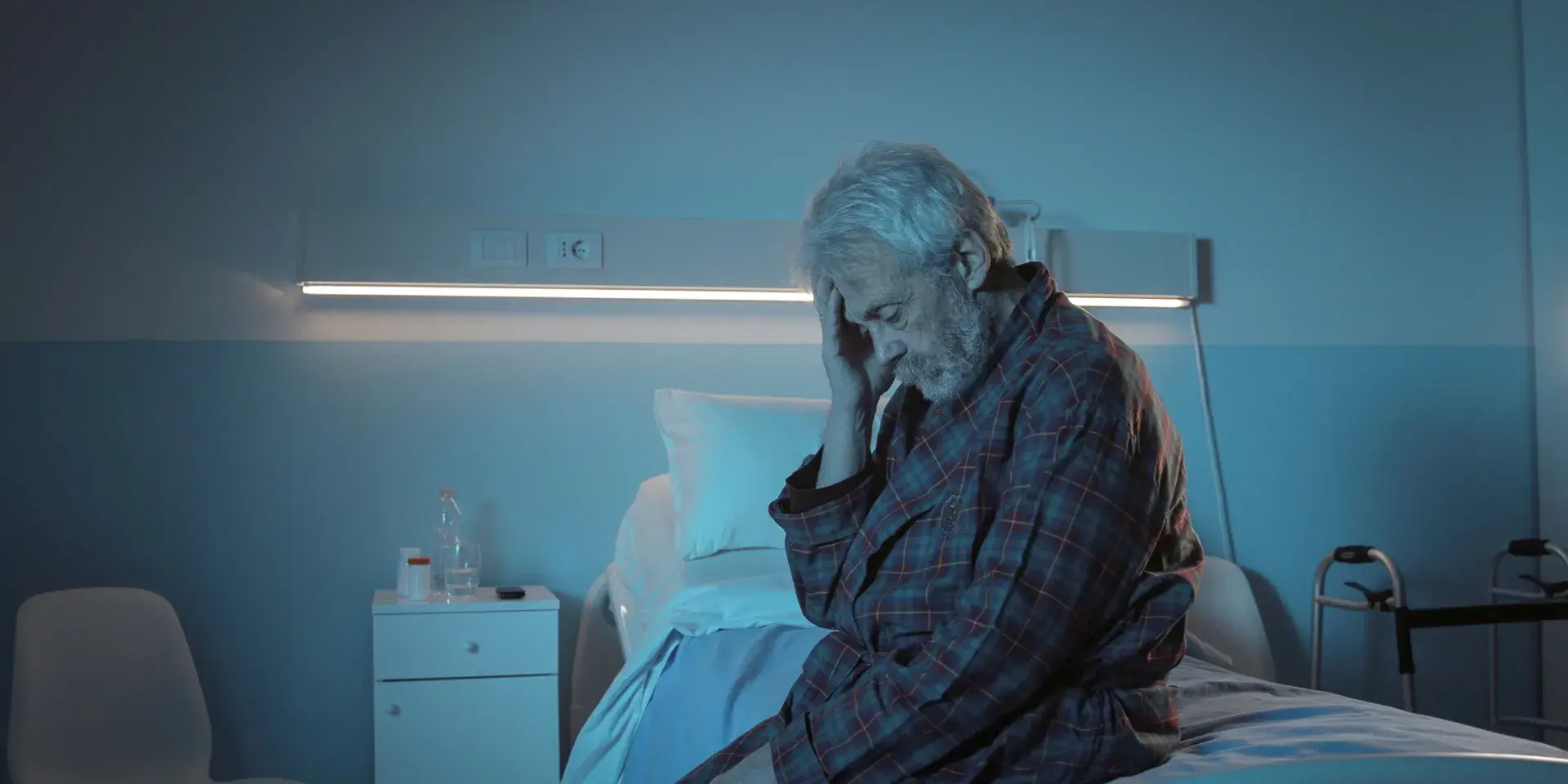 An elderly man sitting in a hospital bed at night and touching his head