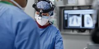 Female surgeon wearing headlamp in operating room. Male and female doctors are wearing blue scrubs.