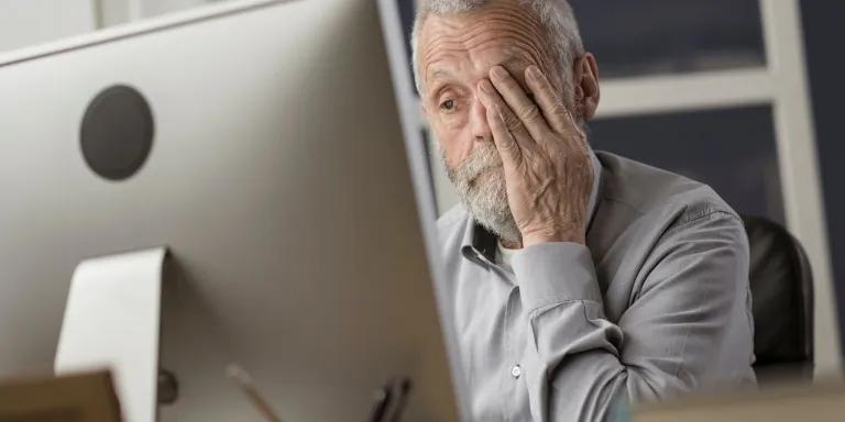 Older man looking tired in front of a computer, rubbing his eyes. 