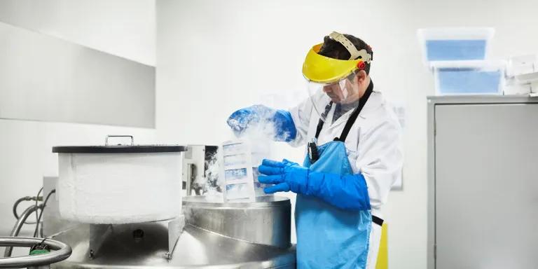 Male scientist examining frozen medicines in freezer. Researcher is working at laboratory. He is wearing protective mask.