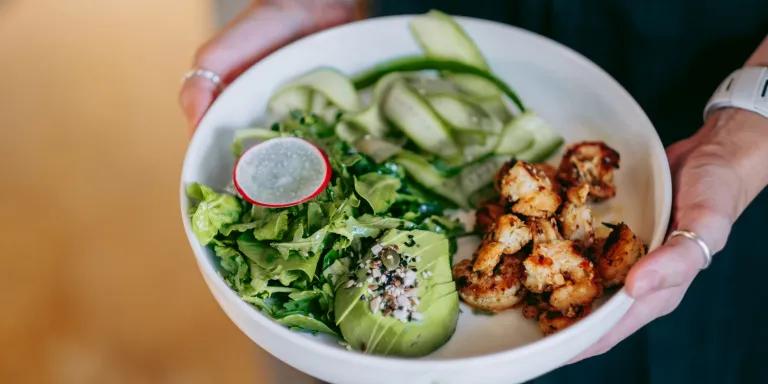  Woman holding white bowl with green salad, avocado, grilled shrimps, cucumber ribbons, radish slice