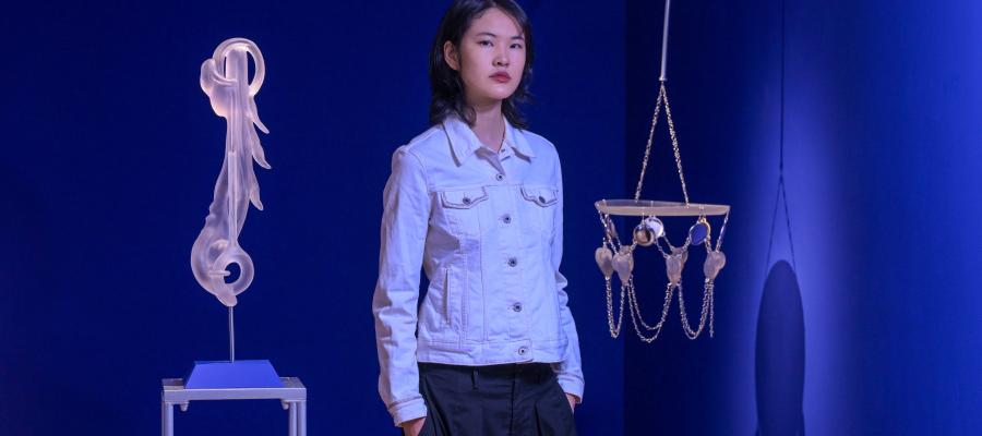 An artist stands in front of their exhibition and holds a piece featuring a woven basket with clay faces.