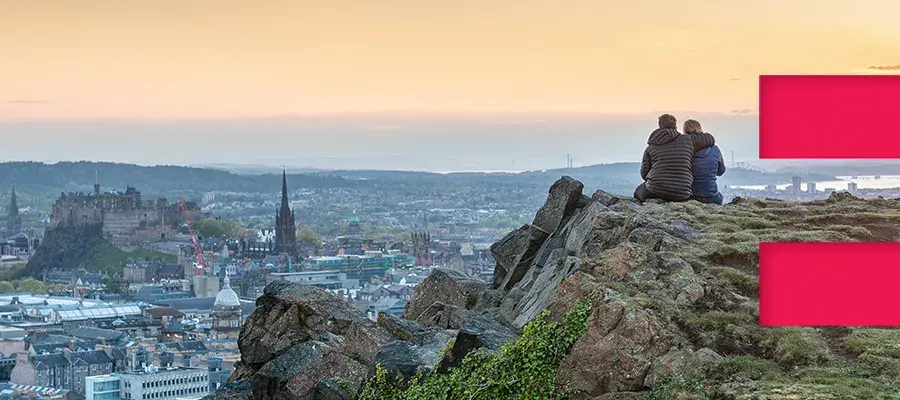 two people sit on the edge of salisbury crags looking out over edinburgh. A lrage red E is on the righthand side of the image