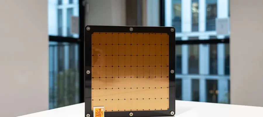 An imager of a wafer-scale AI chip sitting on its side of a white table surface with a postage stamp in front of it. The chip is around the same size as a dinner plate, but is square with a black frame and a gold surface. 