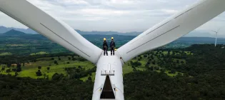 Two people in yellow hard hats stand on top a wind turbine and look at green countryside
