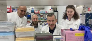 Four members of the REACT Edinburgh team pictured in the lab wearing white lab coats with lab materials in the foreground