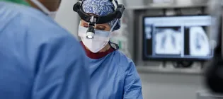 Female surgeon wearing headlamp in operating room. Male and female doctors are wearing blue scrubs.
