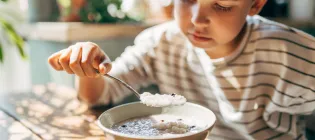 Young boy eating porridge at table near a window 