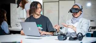 two people sit at a desk. One has VR googles on, the other nakes changes on a laptop