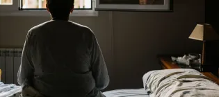 Older woman pictured from behind, sat on a bed looking out of a window