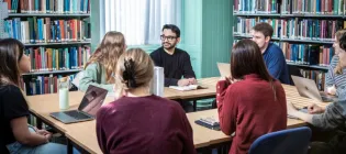 A group of eight students are sat around a rectangular table. Each has a laptop or notebook in front of them and they are engaged in group discussion.