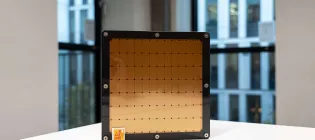 A photograph of a Cerebras AI chip sitting on its side on a white table with a postage stamp in front of it to provide scale. The postage stampe is about 1 per cent the size of the AI chip.