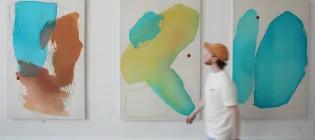 A person walks in front of three paintings at the ECA Graduate Show exhibition