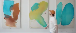A person walks in front of three paintings at the ECA Graduate Show exhibition
