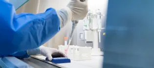 Researcher wearing blue lab coat and gloves uses a pipette to transfer liquid in the lab