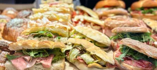 Close-up of multiple rustic sandwiches displayed on a bakery counter, featuring crusty bread filled with a mix of cured meats and salad