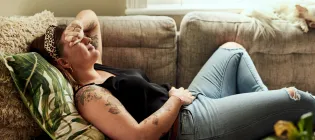 Young woman suffering from stomach cramps on the sofa at home, with hand over her face in pain