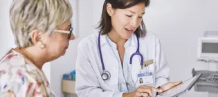 Female doctor explaining electronic test results to female patient at clinic. They are sitting in examination room and the doctor has an ipad. The doctor is wearing a white coat and has a stethoscope around her neck. The patient is wearing a floral shirt and glasses.