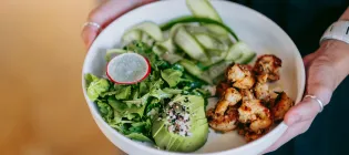  Woman holding white bowl with green salad, avocado, grilled shrimps, cucumber ribbons, radish slice