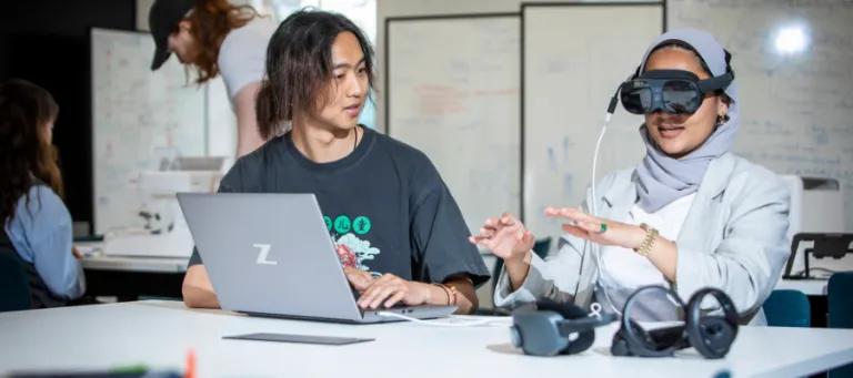 two people sit at a desk. One has VR googles on, the other nakes changes on a laptop