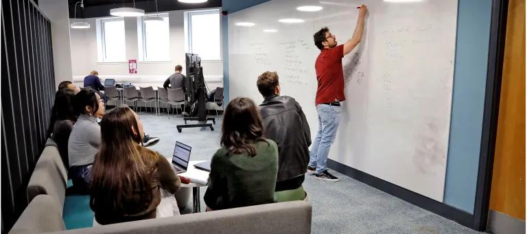 A man in a red t-shirt is standing in front of a large white board and writing on it. Sitting on a sofa in front of him are six people looking at what he's writing. 