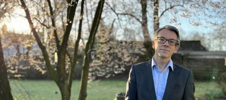 A photograph of Ewen Harrison who has grey hair, glasses and is wearing a suit jacket with a blue shirt underneath. He is standing outside underneath some trees. 
