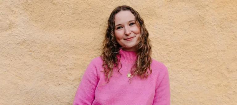 Emma Ahlert - wearing a pink jumper standing in front of a cream textured wall