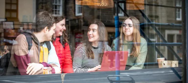 Students photographed through a window. They are sitting down and looking at a laptop.
