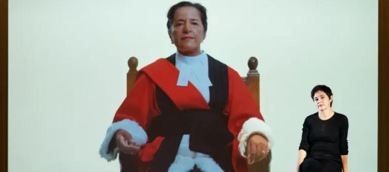 A person in a judicial robe is displayed on a large screen, sitting on a wooden chair. To the right, a second person in casual attire is seated on the floor, with a relaxed posture, looking at the first person.