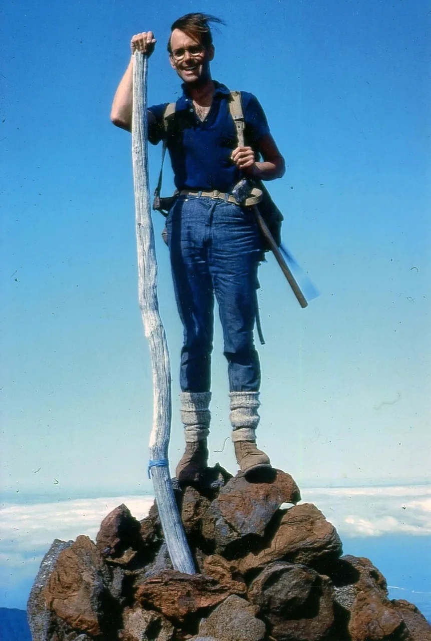 A young Professor Upton stands at the top of a rocky volcano with blue skies behind him