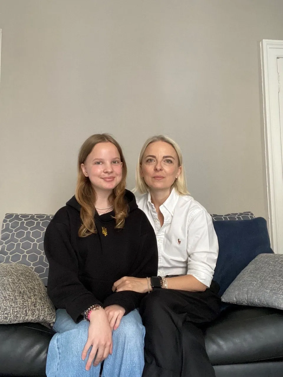 A young blonde woman wearing jeans and a black hoodie sits together with her mother on a black sofa with grey patterned cushions